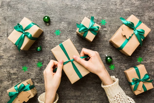 Woman is packing surprise gift for Christmas holidays, sitting at workplace with craft tools, top view. Woman wrapping Christmas gift at colored table.