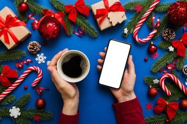 Bir kadının elinde sıcak bir fincan kahveyle akıllı bir telefonu tutması. Renkli arka planda Noel süslemesi.