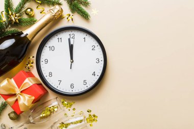 New year celebration concept with a bottle of champagne and two glasses toasting. Christmas gift box, alarm clock and fir tree branch on colored table. Top view Copy space.