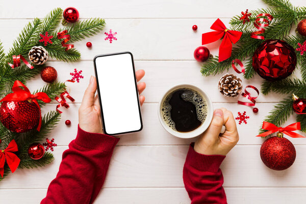 Above view of female hand holding smart phone with hot cup of coffee. Christmas decoration on colored background.