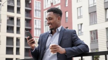 Modern binaların yakınında akıllı telefon kullanarak gülümseyen genç siyah işadamı.