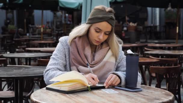 Una mujer en la terraza en un restaurante Chica rubia con ojos azules sentado en la cafetería de la ciudad al aire libre hace notas en un cuaderno de otoño día. La chica usa un diario para escribir ideas. Motivación. — Vídeos de Stock