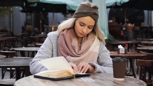 Piękna Blondynka z niebieskimi oczami siedząca w miejskiej kawiarni na świeżym powietrzu robi notatki w jesienny dzień notatnika. Dziewczyna używa pamiętnika, żeby spisywać pomysły. Planowanie kreatywnych pomysłów. Uzasadnienie. — Wideo stockowe