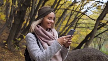 Üzerinde cep telefonu olan mutlu, düşünceli, sarışın bir kadın. Sosyal medyada geziniyor. Golden Autumn Park 'ta akıllı telefonun ekranını kaydırıyor..