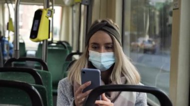 Tıbbi maskeli güzel sarışın kadın toplu taşıma araçlarında cep telefonuyla dinleme mesajları kullanıyor. Tramvay. Sağlık ve güvenlik kavramını, koronavirüs karantinasını, virüs korumasını.