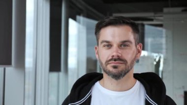 Ofisteki yakışıklı ve serbest çalışan bir işadamının portresi cam bölümlerin arka planında. Gülümseyen adam kameraya beyaz tişört ve siyah kazak giyerek bakıyor. İş için. Başarılı insanlar..