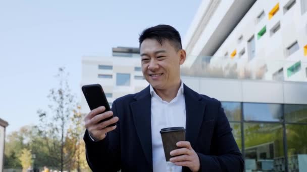 Hombre asiático mensajes de texto en el teléfono inteligente, mientras que de pie en la calle con una taza de café. — Vídeo de stock