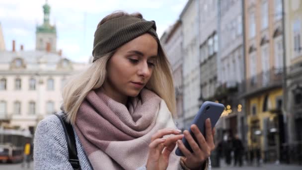Wunderschönes blondes Mädchen mit charmantem Lächeln und blauen Augen, das in der City Street steht, während es mit der neuen Social-Media-App auf dem modernen Smartphone surft. Attraktive Lady tippt Nachricht online. — Stockvideo
