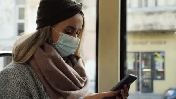 Närbild porträtt av unga smarta kvinnliga student i en medicinsk mask sms:ar på telefonen i en kollektivtrafik. Spårvagnsfönster. Gamla europeiska staden. Karantän COVID-19 i Europa. Coronavirus. — Stockvideo