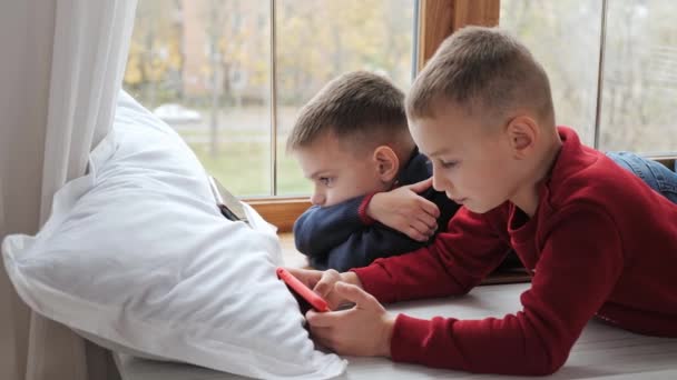 Dois meninos irmãos deitados no parapeito da janela assistindo desenhos animados em telefones celulares. — Vídeo de Stock