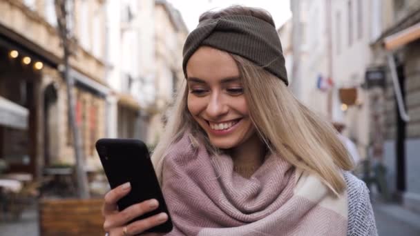 Lächelndes schönes blondes Mädchen mit charmantem Lächeln, das in der City Street steht, während es auf dem modernen Smartphone surft. Attraktive Dame tippt Nachricht, kommuniziert online mit Handy. — Stockvideo