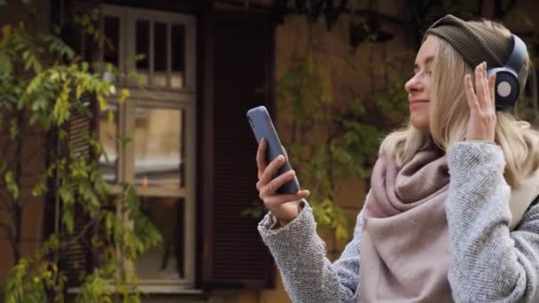 Telefon alkalmazás gyönyörű lány a városi utcákon háttér hallgat zenét vezeték nélküli kék fejhallgató. Boldog nő visel kabát játékosan táncol okostelefon őszi nap. — Stock videók