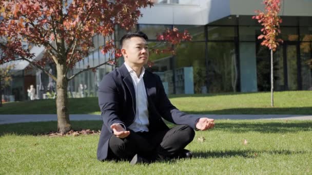 Hombre de negocios asiático meditando y practicando yoga mientras está sentado en la hierba. — Vídeo de stock