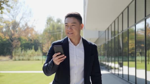 Азиатский успешный бизнесмен ходит с телефоном на открытом воздухе рядом с офисными зданиями. — стоковое видео