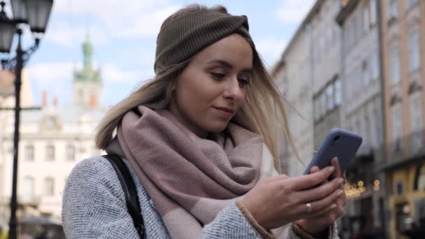 2017 년 11 월 17 일에 확인 함 . positive Beautiful Blonde Girl with Charming Smile Standing in City Street while Browsing Modern Smartphone using Social Media new App. 성공, 스타트업 비즈니스, 온라인 워킹. 위기에서의 성공. — 비디오