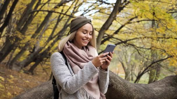 Jonge aantrekkelijke blonde vrouw met behulp van een blauwe smartphone op een achtergrond van vergeelde bomen in de herfst. mooi meisje in een jas schrijft een bericht afluisteren surfen internet op de telefoon in het park. — Stockvideo