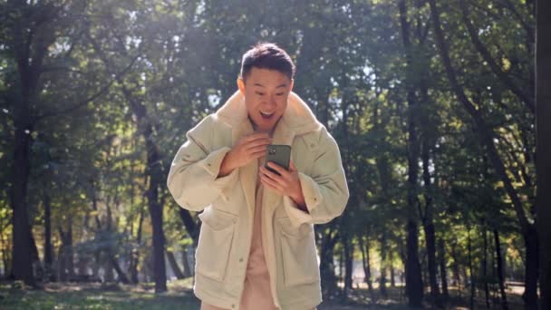 Blogueiro asiático desfrutar de sucesso enquanto olha na tela do smartphone. Notícias positivas. — Vídeo de Stock