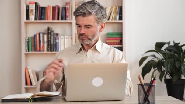 Selbstbewusster erwachsener männlicher Lehrer benutzt Laptop zu Hause und macht sich Notizen am Arbeitsplatz. — Stockvideo