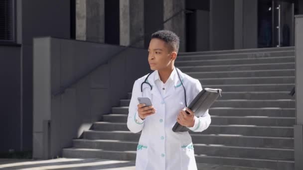 Afrikai-amerikai orvos sétál a kórház közelében az okostelefon internetes alkalmazással. — Stock videók