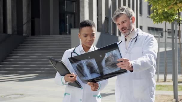 Dwóch profesjonalnych lekarzy analizujących zdjęcie rentgenowskie przy wejściu do szpitala. — Wideo stockowe