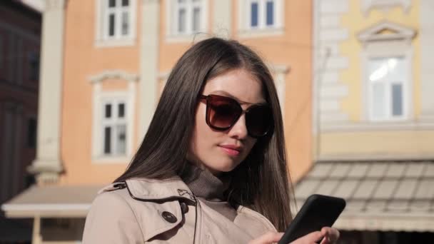 女の子は屋外カメラに笑みを浮かべて町に立っている間、携帯電話でテキストメッセージ. — ストック動画