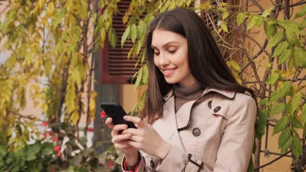 Mensagens de texto feminino feliz no celular enquanto está perto da parede com plantas. — Vídeo de Stock