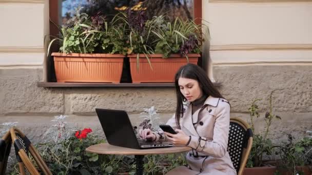 Красивая женщина держит мобильный телефон ввод данных на ноутбук на открытом воздухе. — стоковое видео