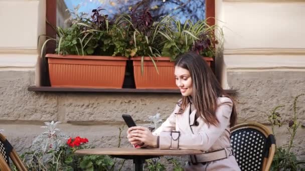Happy girl SMS-y na telefon komórkowy podczas siedzenia na tarasie restauracji. — Wideo stockowe