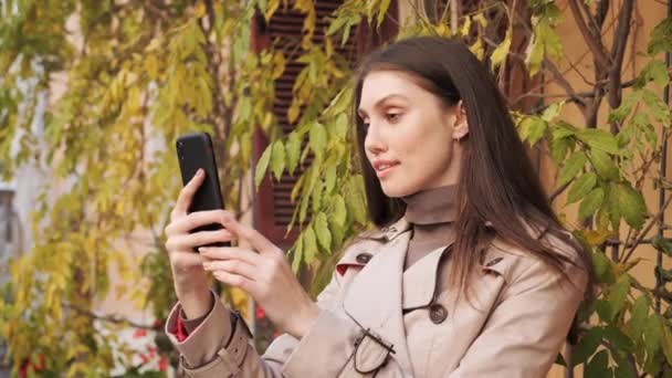 Bela feliz jovem menina vestindo um casaco fazendo um vídeo chamada ao ar livre. — Vídeo de Stock