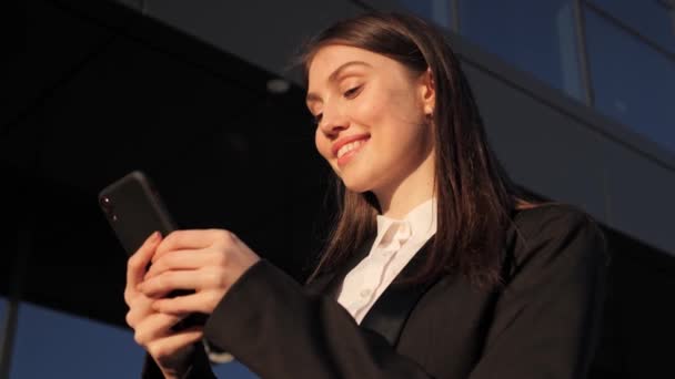 Uśmiechnięta kobieta biznesu Stojąc w pobliżu centrum biurowego przeglądania Nowoczesny smartfon. — Wideo stockowe