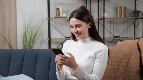 Jolie femme brune assise sur un canapé bleu utilise le sourire du téléphone dans un appartement moderne. — Video