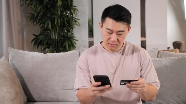 アジアの男の顧客を保持クレジットカードスマートフォンに座ってソファに自宅. — ストック動画