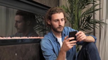 Sakallı neşeli adam telefon uygulaması kullanarak şöminenin yanında oturuyor..