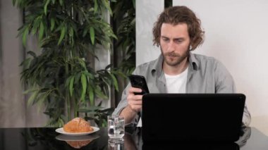 Bilgisayarlı akıllı telefon kullanan bir adam ofis masasında oturmuş mesajlar yolluyor..