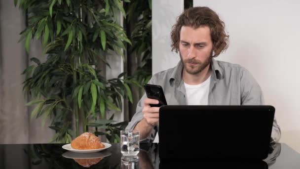 自宅のオフィスのテキストメッセージでテーブルに座ってノートパソコンのスマートフォンを使用して男. — ストック動画