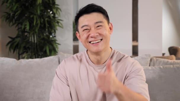 Feliz asiático alegre guapo hombre mostrando pulgar arriba signo sentado en sofá en casa. — Vídeos de Stock