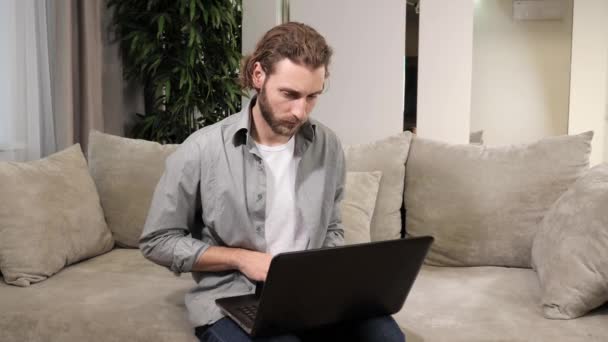 Hombre guapo trabajando en el ordenador portátil mientras está sentado en el sofá en casa. — Vídeo de stock
