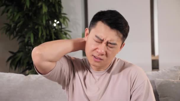 Asiático hombre sentado en sofá en casa exceso de trabajo cansado, experiencias dolor en el cuello. — Vídeos de Stock