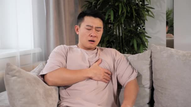 Nemocné srdce Problémy s dýcháním, bolest na hrudi, asijský muž sedí na gauči doma. — Stock video