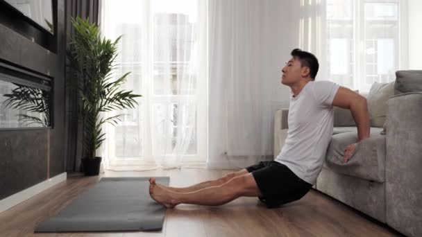 Athletic asian man doing exercises squeezes leaning on the sofa at home. — Stock Video