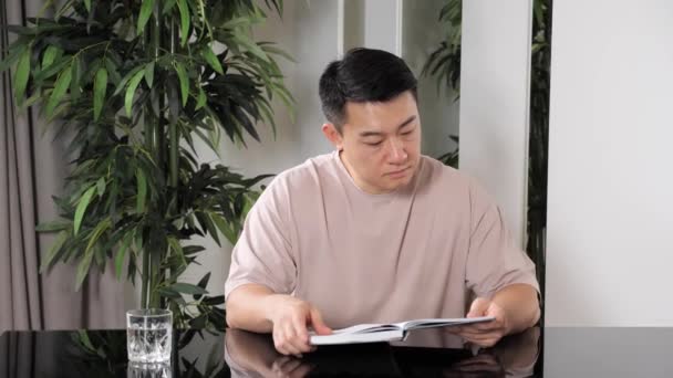 Ein hübscher asiatischer Mann in lässiger Kleidung liest zu Hause ein Buch. — Stockvideo