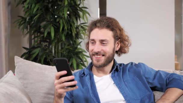 Hombre joven guapo usando mensajes de texto de teléfonos inteligentes sentado en el sofá. — Vídeos de Stock