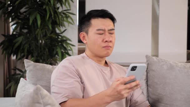Azji człowiek siedzi na kanapie używa telefonu i ma ostry ból zęba problem stomatologiczny. — Wideo stockowe