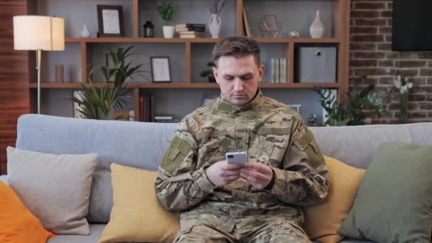 Un soldat concentré en uniforme militaire est assis sur un canapé à la maison et feuillette les photos après la guerre. Soldat masculin défilant sur l'écran de la messagerie smartphone avec sa petite amie ou sa femme. — Video