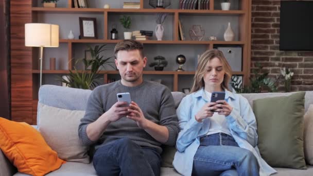 Glada unga par sitter på soffan surfa sociala medier internet med mobiltelefon på mysiga soffan njuter helger tillsammans. Pojkvännen och flickvännen är hemma. Modern teknik — Stockvideo
