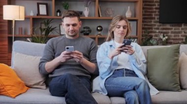 Çift kanepede birlikte oturuyor ve telefonlarını kullanıyor, genç çift yaşam tarzı, boş zaman aktiviteleri. Kız, sosyal ağlardan erkek arkadaşına fotoğraflar gösteriyor. Cep telefonlarına bağımlılık. 
