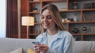 Evde akıllı telefon cihazıyla cep telefonu tutan mutlu güzel kız. Gülümseyen sarışın kadın blogcu yeni sosyal medya aboneliği, internetten satın alma, uygulamalarla ürünler sipariş etme. 