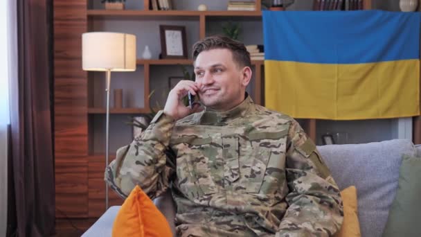 A férfi jóképű katona katonai egyenruhában otthon ül a kanapén, és a családjával vagy a barátaival beszélget. Ukrán zászló a háttérben. Katonai akciók Ukrajnában. Szabad demokratikus emberek, vessenek véget a háborúnak!. — Stock videók