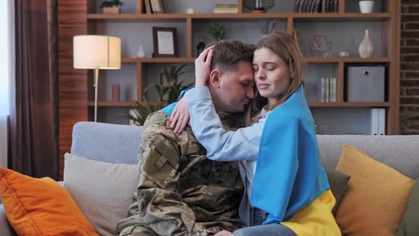 Soldado masculino em uniforme militar e sua menina envolto na bandeira da Ucrânia abraçando em casa no sofá. hostilidades militares. Mobilização da população. Os russos atacam a Ucrânia. Parem a guerra.. — Vídeo de Stock