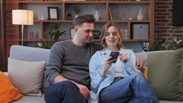 Pareja de la familia relajarse en el sofá utilizando el teléfono móvil inteligente para las compras en línea. Feliz pareja milenaria usando divertidas aplicaciones sonrientes hombre y mujer relajándose en casa, divirtiéndose en las redes sociales en línea. — Vídeos de Stock